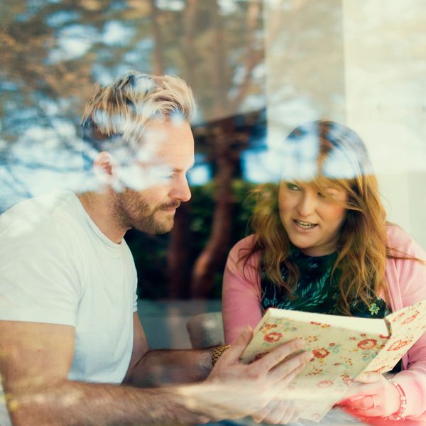 A man and woman discuss a floral-patterned book indoors through a window reflection.