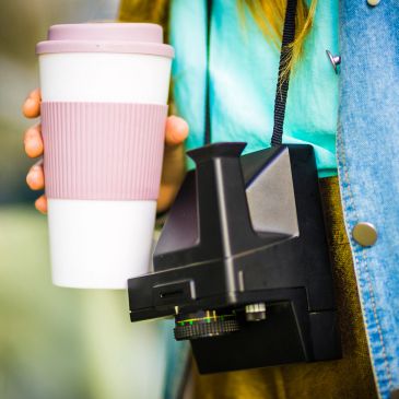 Person holding a coffee cup with a camera hanging from their neck.