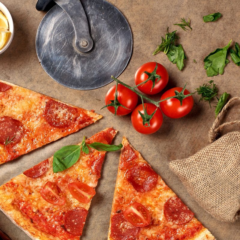 Flatlay or top view. Pepperoni pizza slices on craft bakers paper. Fresh tomatoes, parsey, circle knife and dill