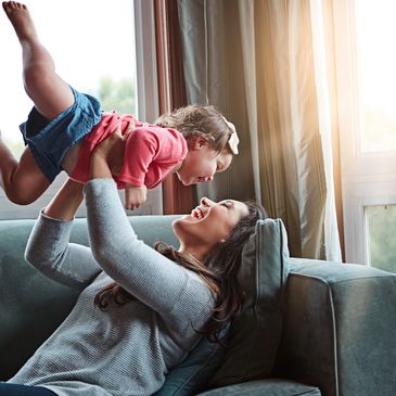 Mother joyfully lifting her laughing toddler on a couch by the window.