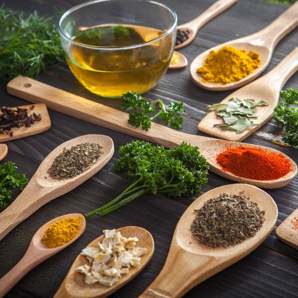 Variety of spices and herbs arranged on wooden spoons with a glass bowl of oil.