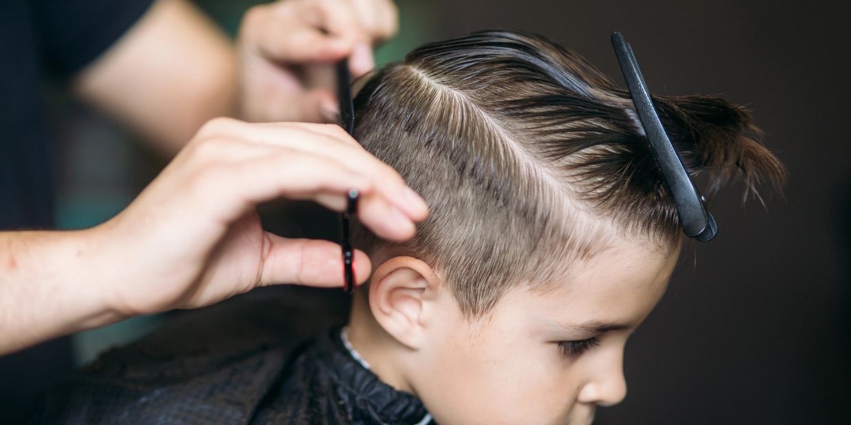 A stylist using a comb and scissors to cut a young boy's hair, styled with a wet side parting.