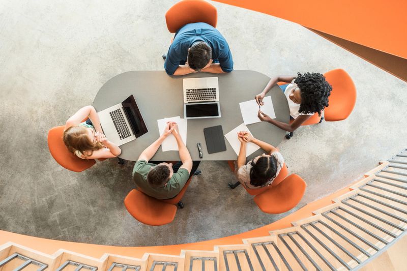 Young women and men sitting on orange chairs in discussion with laptops and notes on table