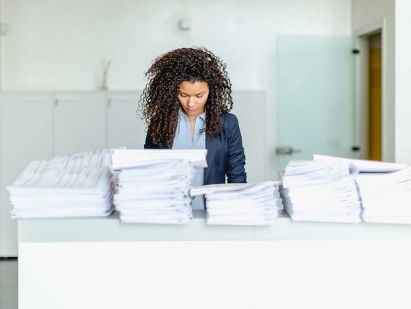 Woman overwhelmed by large stacks of paperwork at office desk.