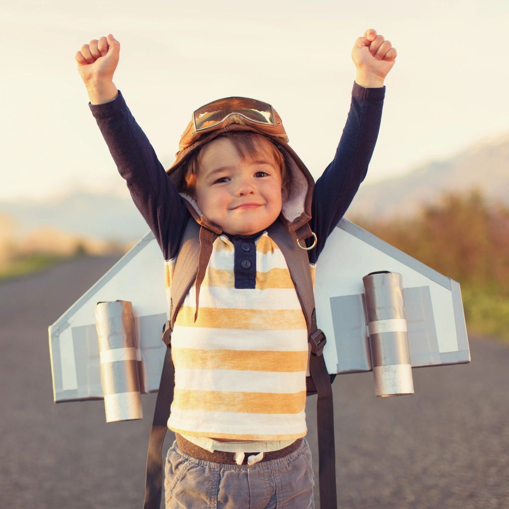 Young boy with toy airplane wings raises arms in excitement outdoors.