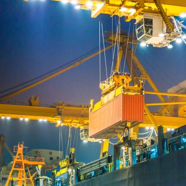 A large crane lifts a shipping container at a brightly lit port at night.
