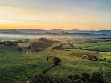 Sunrise over misty rolling hills and farmland with scattered trees and houses.