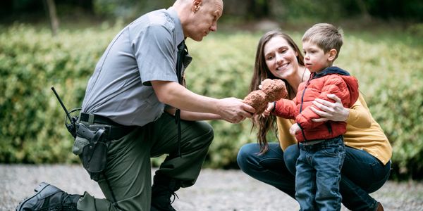 Park ranger giving a teddy bear to a little boy while his mother watches.