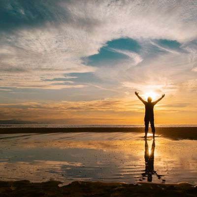 Person standing with arms raised at sunset on the beach.