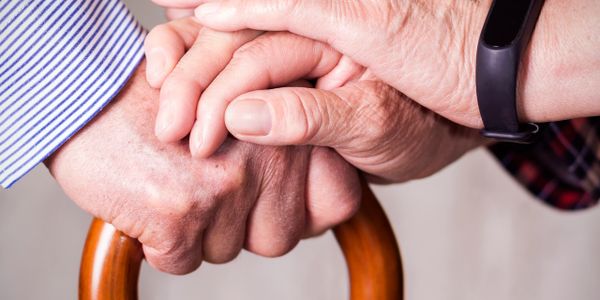 Two elderly hands clasped over a wooden cane, symbolizing support and care.