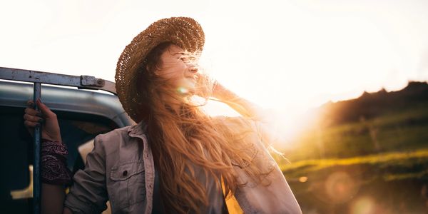 Woman enjoying sunlight with hair blowing, wearing a straw hat and casual jacket.