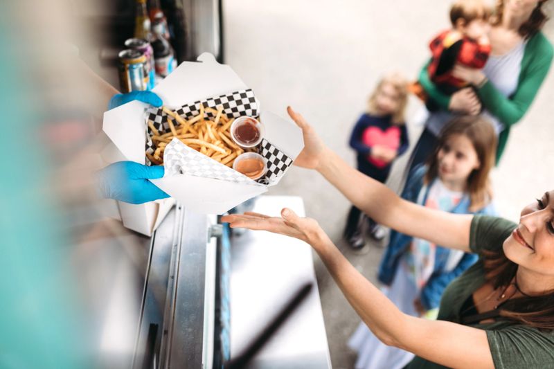 A young family group shares fun food and memories at a local food cart, serving delicious handmade sandwiches and snacks.  One of the women smiles as she receives her order of gourmet french fries with special dipping sauce.