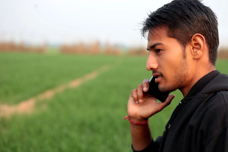 Young men of Asian ethnicity standing in the field of wheat crop during winter season & talking on his smart phone. He is looking happy while talking on mobile phone.