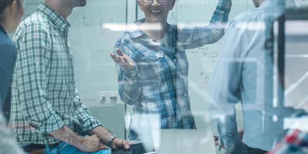 A woman leads a meeting with colleagues in front of a whiteboard filled with diagrams.