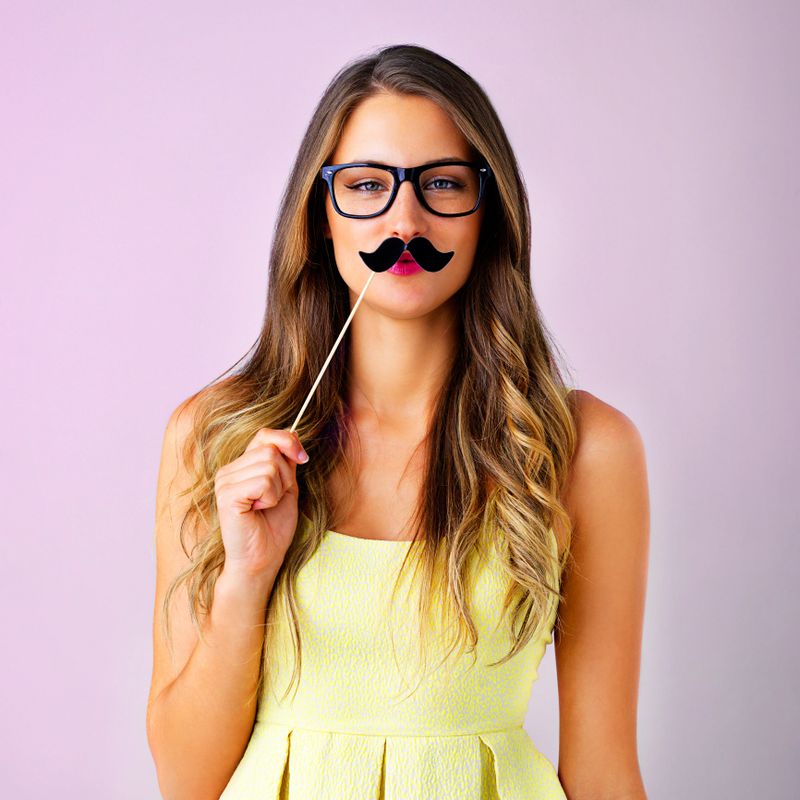 Studio shot of a young woman holding a mustache prop to her face against a pink background