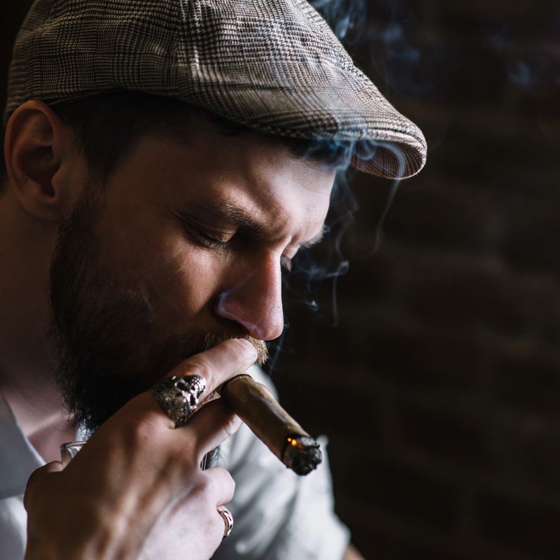 A young bearded man dressed in a vintage style smokes a cigar in a pub, close-up