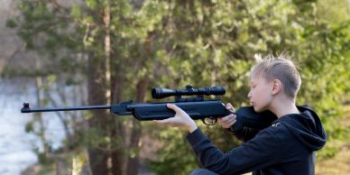 Young boy aiming a scoped rifle outdoors.