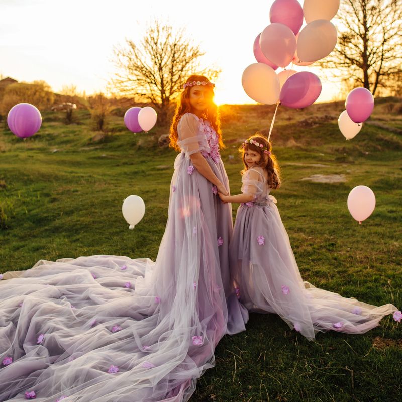 Young beautiful red-haired mother with her daughter, dressed in luxurious purple dresses, walking with balloons in nature in the sun