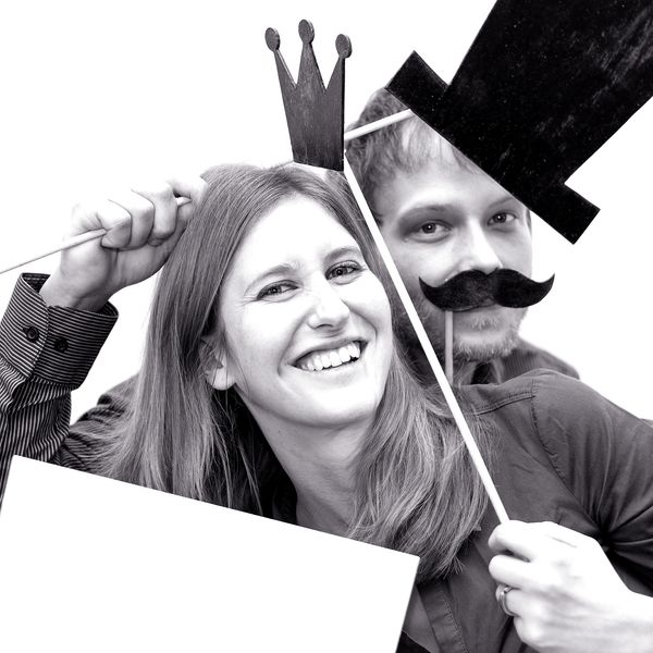 Two people smiling, one holding a stick, the other wearing fake mustache and hat.