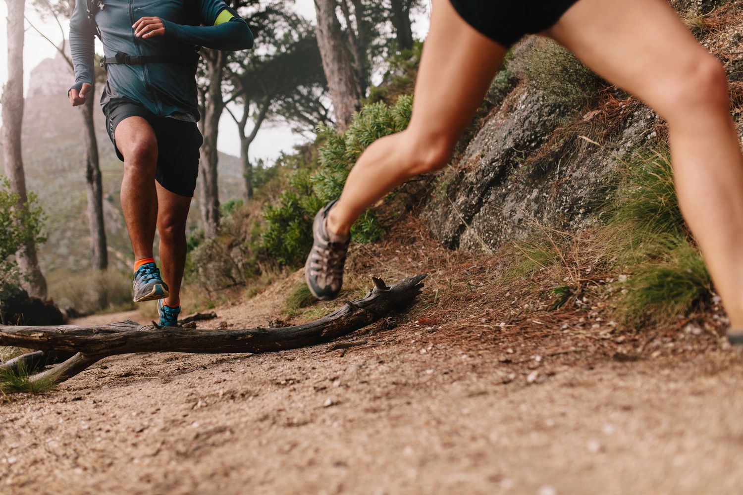 Two people trail running outdoors, jumping over a fallen log on a dirt path.