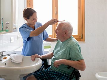 Caregiver gently combs elderly man's hair in a bright bathroom.