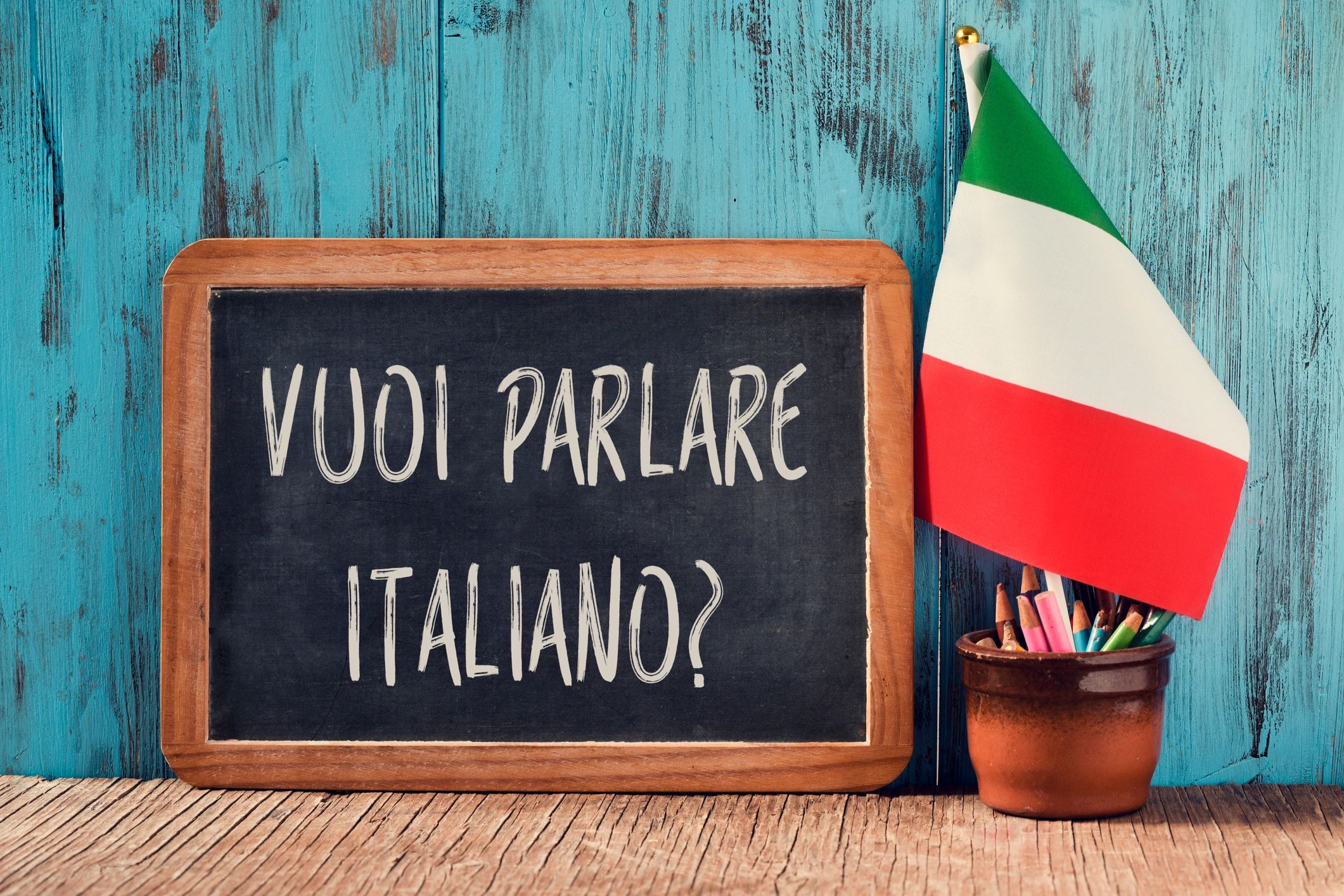 Blackboard with 'Vuoi parlare italiano?' next to an Italian flag and colored pencils.
