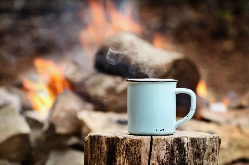 Blue enamel cup of hot steaming coffee sitting on an old log by an outdoor campfire. Extreme shallow depth of field with selective focus on mug.