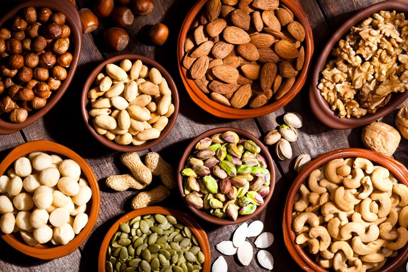 Top view of a rustic wood table filled with a large assortment of nuts like pistachios, hazelnut, pine nut, almonds, pumpkin seeds, peanuts, cashew and walnuts. Some nuts are in brown bowls and others are placed directly on the table. Predominant color is brown.