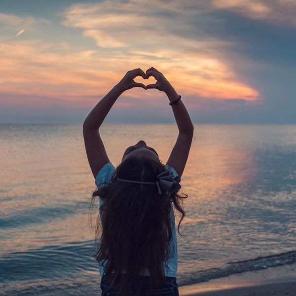 Girl making a heart shape with hands at sunset by the sea.