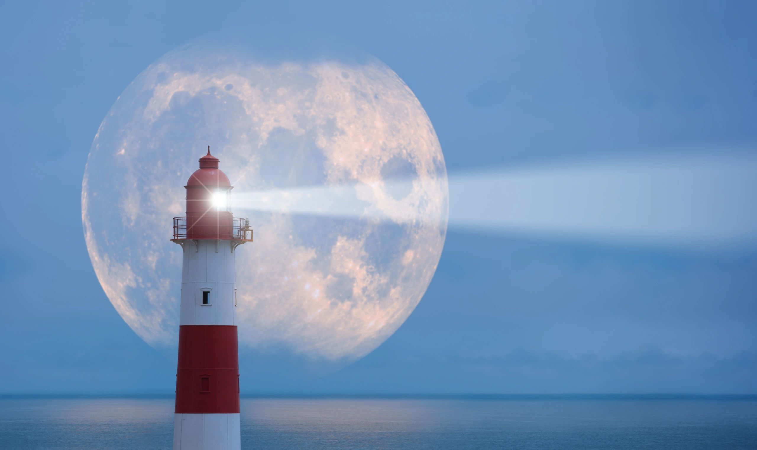 Lighthouse beam shining with a giant full moon in the background over calm sea.