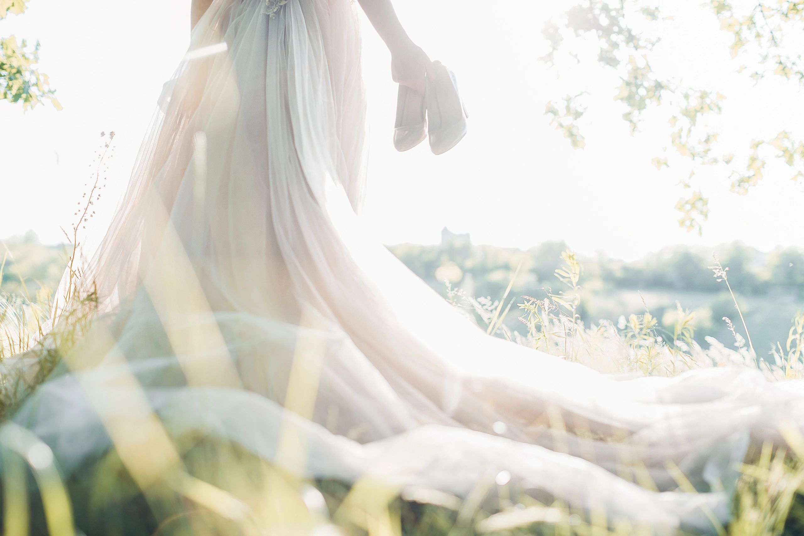 Bride holding shoes, standing in a sunlit field with flowing wedding gown.