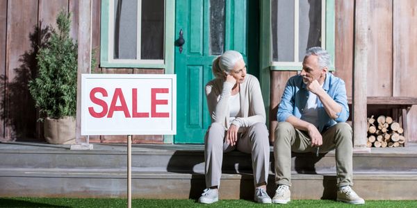 Elderly couple sitting on porch steps beside a sale sign in front of a house.