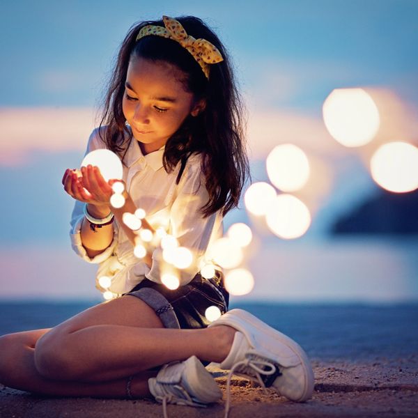 A girl sitting on the beach holding glowing string lights at twilight.