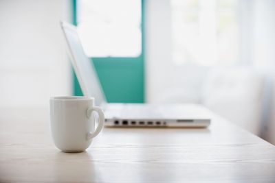 White coffee mug on a wooden table with a laptop blurred in the background.