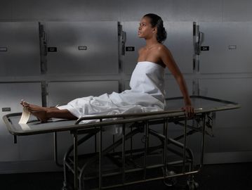 Woman wrapped in white cloth sitting on a morgue stretcher.