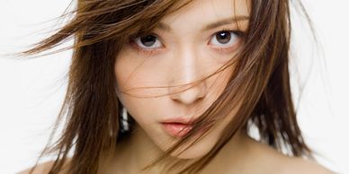 A close-up of an East Asian woman with long brown hair, strands blowing across her face.