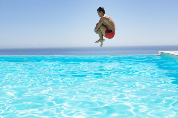 Man in red swim trunks jumping into a bright blue pool with ocean in background.