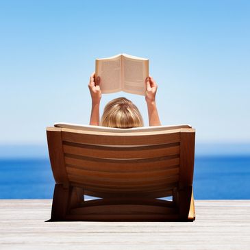 Person relaxing on a wooden lounge chair by the sea, reading a book.