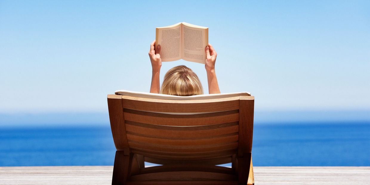 Person reading a book on a wooden lounge chair by the sea.
