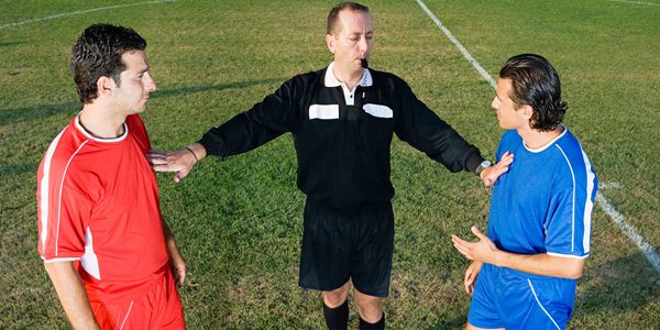 Referee mediating between two soccer players in red and blue uniforms on the field.