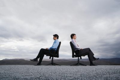 Two businessmen sitting back-to-back on office chairs outdoors.