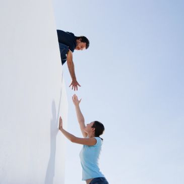 A man reaching down to help a woman up against a white wall under a clear sky.