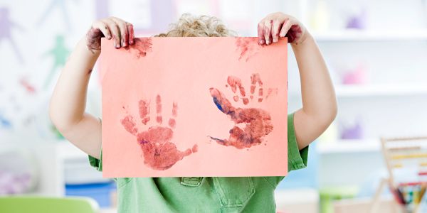 Child holding paper with red handprints, hiding their face.