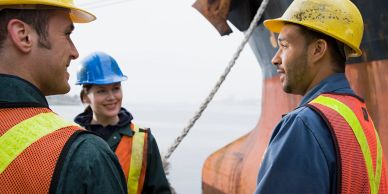 Three dock workers in safety gear talk near a ship at the harbor.