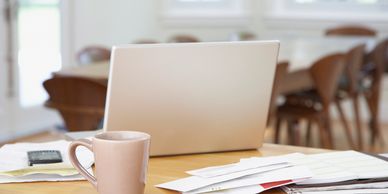 A laptop, coffee mug, and scattered papers on a wooden table in a bright room.