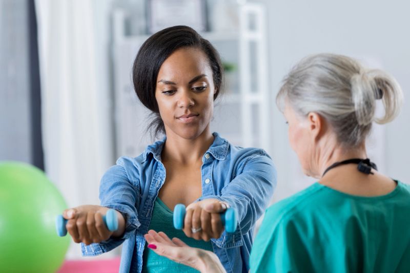 Mid adult African American woman lifts hand weights in physical therapy session. A senior physical therapist is teacher her the proper technique for lifting the weights.