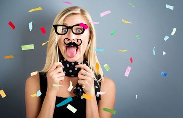 Woman with playful disguise glasses and mustache sticks out tongue amid colorful confetti.