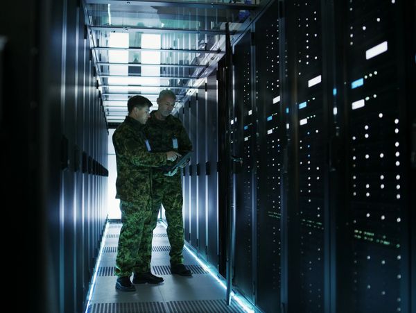 Two soldiers in camouflage uniforms inspecting servers in a data center.
