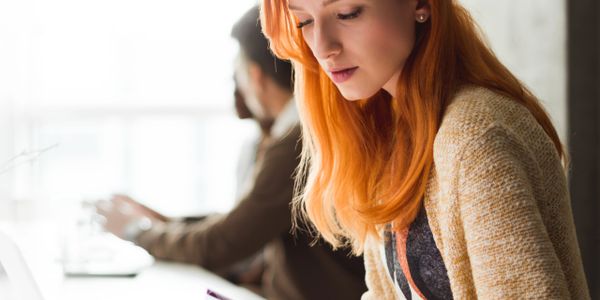 A focused woman with red hair working at a desk with a blurred man in the background.