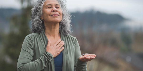 A woman practices mindfulness outdoors with eyes closed and hand on chest.